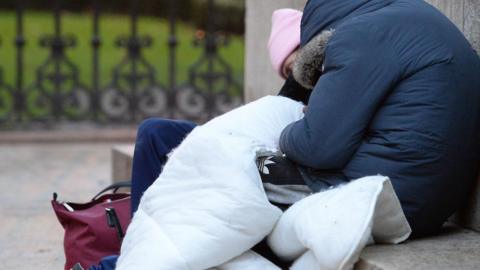 A homeless person sat outside with all of their belongings, including a bag and a duvet without a duvet cover; they are wearing a pink hat, navy coat and navy adidas joggers.