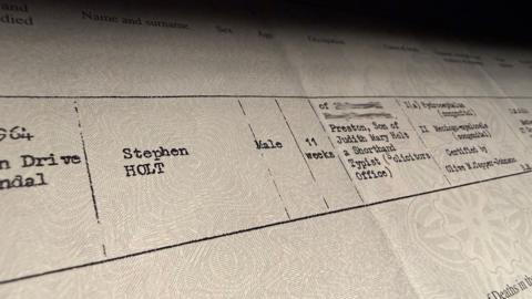 Close-up of an official UK death certificate from the General Register Office, showing typed details and headings, including registration district and cause of death, photographed under soft lighting.