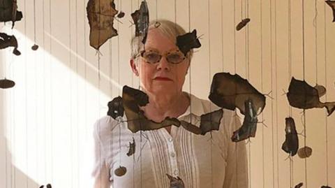 Sara Davidmann wearing a white shirt, glasses and black trousers standing behind burnt photo fragments and stones hanging from the ceiling on strings