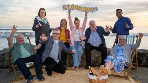 A group of men and women celebrating on a beach with a £1,000,000 sign 