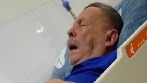 Michael Blake lying on a gurney in hospital while wearing a blue top. He has his eyes closed and his mouth open.