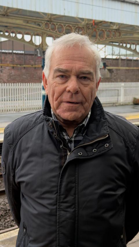 Ken Harper is standing on a railway station platform and looking directly into the camera. He has short grey hair and is wearing a dark blue coat. Behind him, the station has circular designs on the arches holding the roof up.