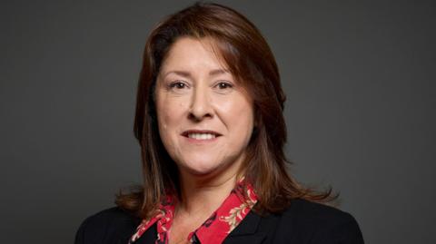 Michelle Scrogham has long brown hair and is wearing a black jacket. She is posing for an official portrait.