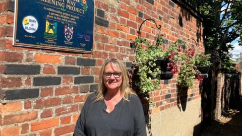 Cait O'Reilly is standing by a brick wall with a plaque on it highlighting the Blombish alleyway community gardening project. There are hanging baskets in the background. Cait is wearing a black top and glasses and is smiling. 