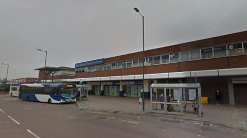 A blue and white single-decker bus is stopped at a bus station which has a row of shops behind. There are people standing at a bus shelter on the right hand side of the image. Three tall lampposts can be seen. The sky is grey and cloudy.