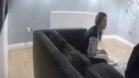 Kerri Pegg sits on a dark sofa with grey walls covered in bird stencils and laminate flooring