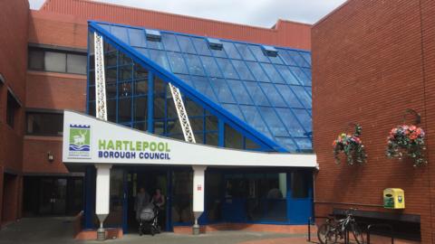 Front of Hartlepool Borough Council HQ, showing the main entrance, a blue metal and glass atrium, with brick walls to the side, the right hand one featuring two hanging baskets.