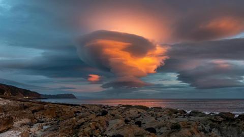 An image of the waters edge at some rocks on the coastline. In the sky is a cone shaped cloud bathed in the soft orange sunlight of the morning sunrise.