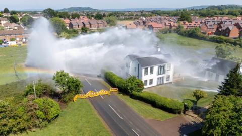 A drone shot of the burst water pipe in Matson, it shows a large torrent of water shooting from a field, across a two lane road and directly onto a large house. The road has been cordoned off with yellow fencing.