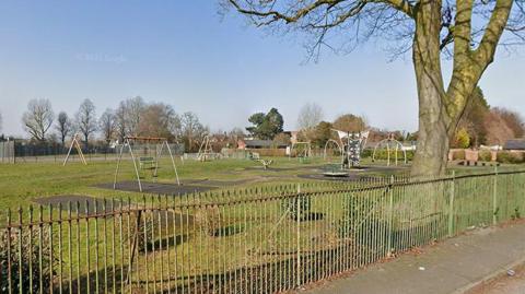 A fenced off play park with swings and climbing apparatus with a large tree to the right on a clear day.
