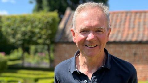 Dr Paul Everden a man who is standing outside in front of green hedges. He is looking directly at the camera and smiling and is wearing a navy polo shirt.