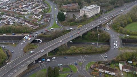 An aerial view of Manadon Roundabout which shows one large road passing over the roundabout with cars travelling around it. 