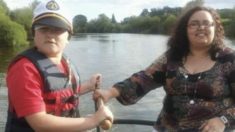 Tomas Cozens as a young boy with his mum Jo. They are on a boat on a river and are holding a wooden handle that steers the boat. Tomas is wearing a red t-shirt and a black and red life jacket with a white, Navy-style captain's hat. His mum has curly dark hair and rectangle-shaped glasses with a dark, patterned long-sleeved top on. The river and trees are visible in the background 