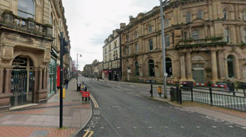 Botchergate in Carlisle. It is a city centre high street lined with old, ornate buildings. There is a pedestrian junction with traffic lights.
