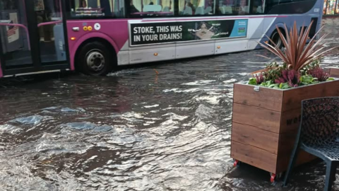 A bus is seen driving through flood water in a town centre. In the foreground is a raised flowerbed and a plastic chair outside a bar.