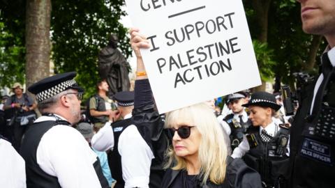 Metropolitan Police officers remove people from a protest in central London in support of Palestine Action. A woman wearing sunglasses with white hair holds a sign saying "I support Palestine Action". 