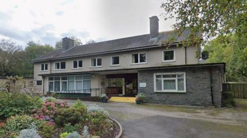 A long 1970s style building made of concrete sits at the top of a short curving drive. It is two storeys and has several windows with shrubs to the left hand side of the drive and trees behind. 