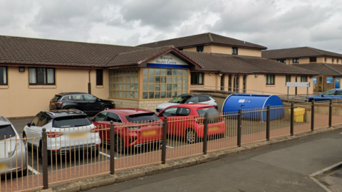 A beige coloured building with several cars parked in front of it. A sign above the entrance reads Blantyre Health Centre.