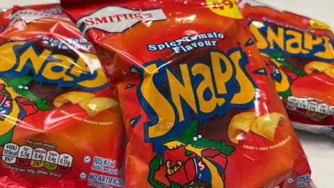 Three crisp packets laid on a table that are all the same. It is a red packet with 'Snaps' written in orange and blue font with a green animal wearing a blue jacket and holding up a bag of crisps and thumbs up. There are rectangular shaped curved crisps pictured on the front that say 'crisp and light potato snacks'. It says Smiths at the top and 49p.