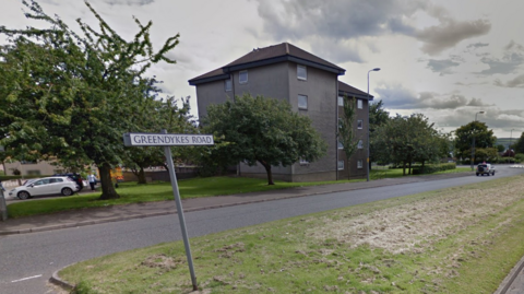 A google view of Greendykes Road showing a street sign in the middle of the road with a four-storey block of flats in the background