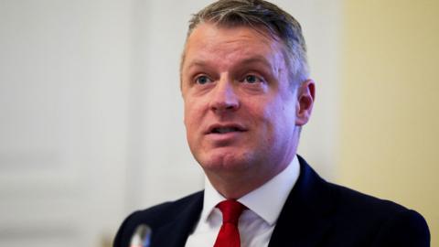 Luke Pollard, a man with grey hair in a dark suit, white shirt and red tie stands in front of a small lectern microphone.