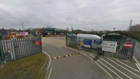 An exterior view of a Hampshire tip with metal fencing, an open gate and waste containers in the background beyond a large parking area