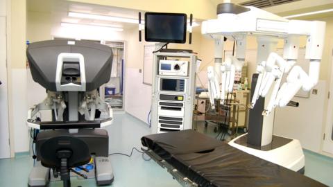 A hospital theatre with a robot with several "arms" above a surgery bed. There are a number of other machines in the room, as well as medicines and cleaning supplies. 