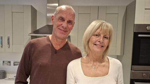 A man and woman are standing in a kitchen. The man is wearing a brown jumper, while the. woman is wearing a cream-coloured top. They are both smiling for the photo.