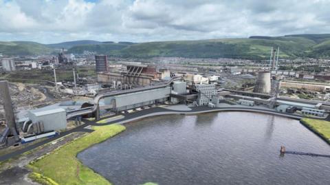 A computer-generated image shows the sprawling site of the steelworks in Port Talbot. A long grey structure represents the new electric arc furnace on the banks of a small lake. It is surrounded by older steel infrastructure including buildings and chimneys, while the terraced houses of Port Talbot and green hills can be seen in the background. 