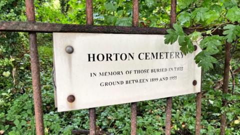 a plaque fixed to a metal railing, inscribed with the words "HORTON CEMETERY in memory of those buried in this ground between 1899 and 1955"