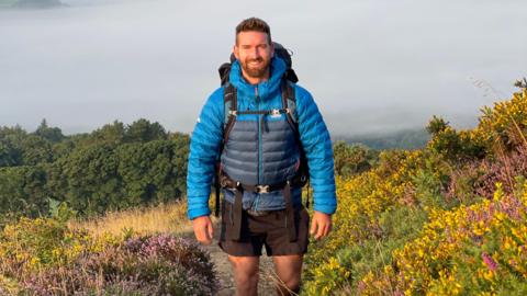 Sam Culley wearing black shorts, a blue puffer jacket and a large backpack. He has short brown hair and a beard and smiling at the camera, standing on a pathway at the top of a tall hill surrounded by heather, flowers and gorse bushes. Behind him is light grey low fog obscuring the view of the landscape.