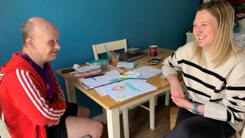 Support worker Kim Roberts sat by a table with paintbrushes, paper and paints, opposite a man wearing a red Adidas hoody