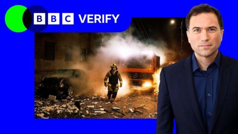 A firefighter walks through debris. A flaming truck can be seen in te background and the man is wearing protective gear. Imposed over the image is the colours of BBC Verify and Ros Atkins. 