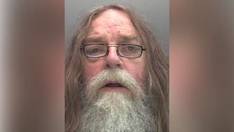 A police custody photo of Douglas Bowyer. He has long brown hair and a long grey moustache and beard. He is wearing black-rimmed, rectangular glasses. The wall behind him is grey.
