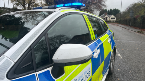 The side of a white, blue and yellow police car with its blue lights turned on. It is parked on a residential street with a cream coloured house in the background. 