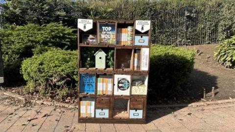 A bookcase promoting the Top of the Wold play standing on a path with greenery in the background, There is a bird box and and a number of brightly-coloured containers on it.