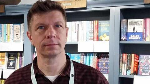 A man in a bookshop wearing a red flannel patterned shirt.