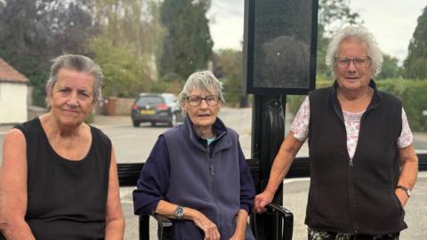Three women in their seventies are standing in front of a black bus stop