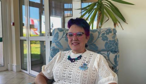 A woman with short, purple hair sat in a blue and white chair. She is wearing a white, short-sleeved knitted jumper with a blue necklace