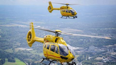 Two yellow helicopters from the East Anglian Air Ambulance flying over some fields, trees, homes and tall buildings.
