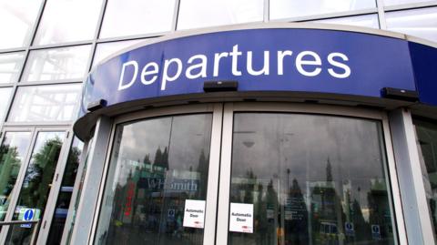 Double automatic doors at the departures entrance of Bristol Airport. There is a big blue sign which says 'departures' above the door.