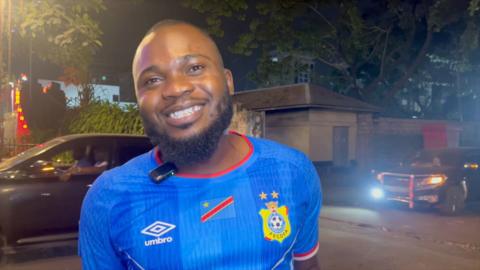 A man wearing a DR Congo jersey smiles to the camera from the streets of Kinshasa
