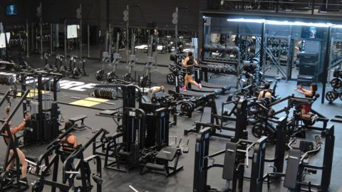 Kulture gym pictured from a high platform inside the warehouse. It is a large open room which has been painted completely black, with lots of gym equipment covering the black mat floors. There are a few people dotted around using the equipment, getting water or lifting weights.