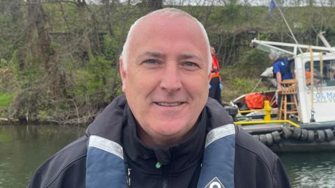 A man with short, white hair wearing a black coat and a blue and black life jacket.