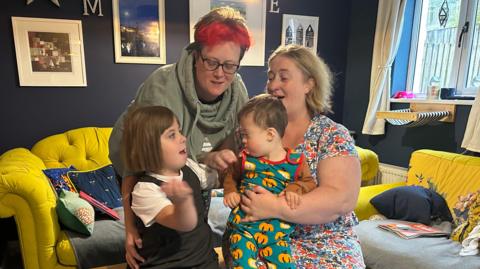 A family of four in their home all looking directly at the camera smiling. There are two adults and two children. They are in their living room which has bright yellow sofas and dark blue walls with lots of picture frames on the wall. 