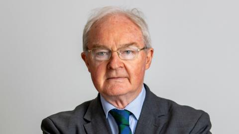 Declan Morgan is pictured against a grey background. He has short grey hair and is wearing silver rimmed glasses. He's dressed in a dark coloured suit, blue shirt and a blue and green tie. 