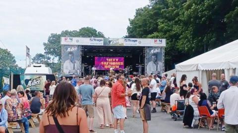 A view of the main stage at the festival showing crowds of people standing and sitting around festival tents.