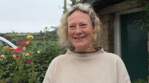 A women with sort curly blonde hair looking smiling and looking just past the camera. She is wearing a light jumper with a black collar. There is a hedge with some flowers over her left hand side shoulder and a black garage door over her right.