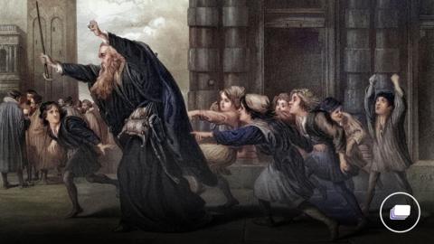 A picture of Shylock from The Merchant of Venice with his arms up holding a stick. There are a group of people behind him. 