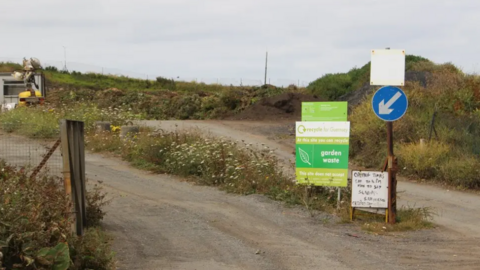 The entrance of a garden waste centre. On the right are two signs. The one of the left is green and there is a smaller white one on the right. There is also a road sign above that. In the background is grass and flowers with a road at the centre.